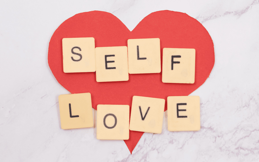 Scrabble tiles spelling "self love" on a red heart-shaped background.