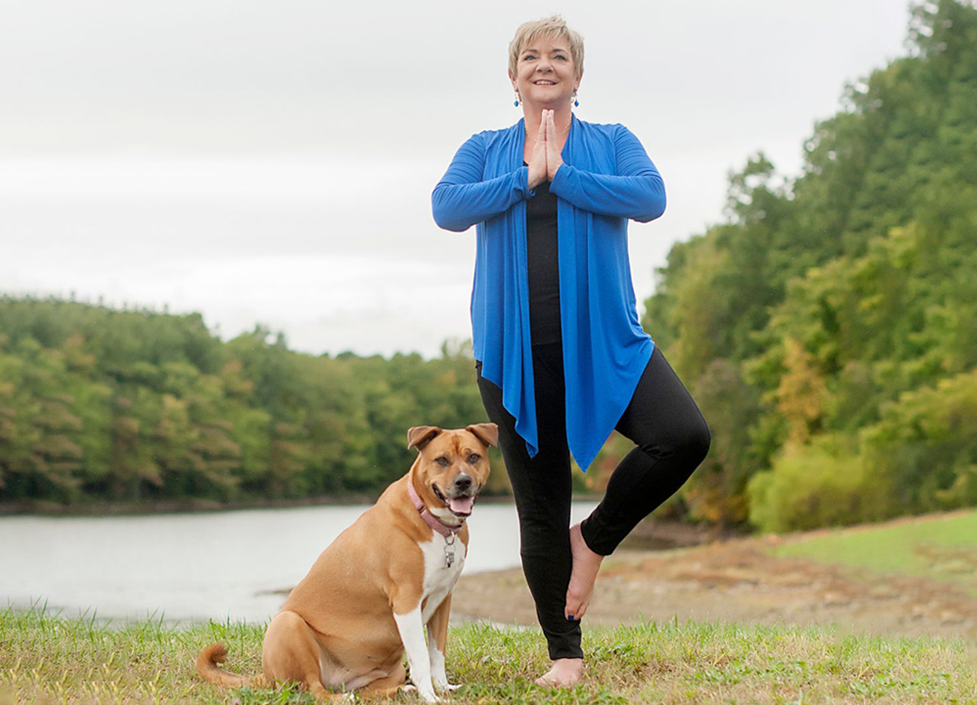 A woman performing yoga next to a sitting dog by a lakeside.