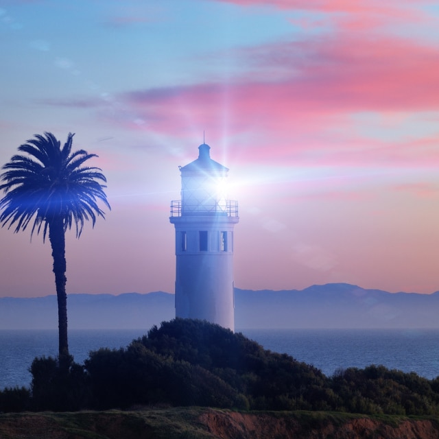 A lighthouse with its beacon shining at dusk beside a palm tree under a colorful sky.