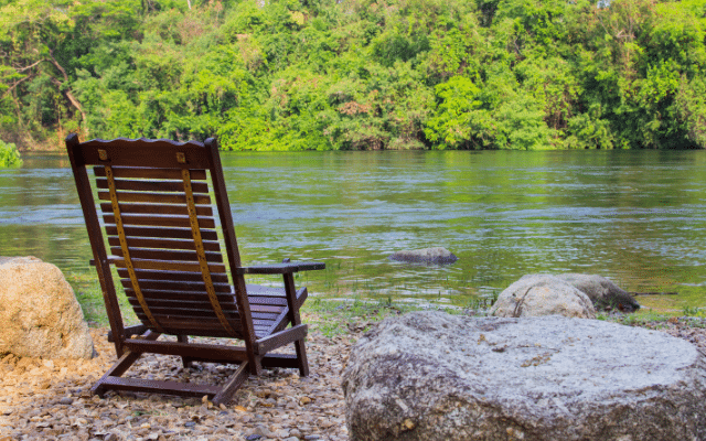 A solitary wooden rocking chair by a tranquil river with lush greenery in the background.