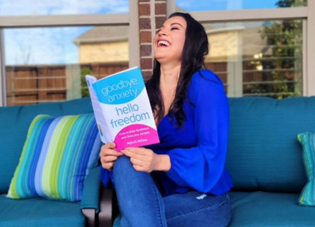 A woman laughs while holding a book titled "goodbye anxiety, hello freedom" on a blue couch outdoors.