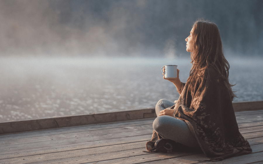 Woman wrapped in a blanket enjoying a warm beverage by the lakeside in the morning mist.