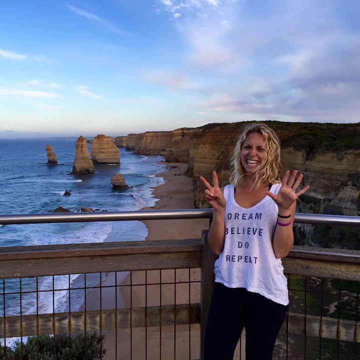 A smiling person posing with a peace sign in front of the twelve apostles rock formations in australia.
