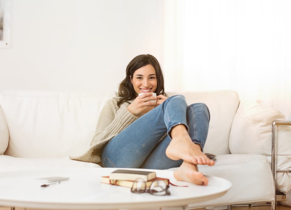 Woman relaxing on a sofa with a cup of coffee, books, and glasses on the table in front of her.