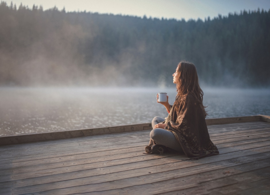A person wrapped in a blanket sitting on a wooden dock by a misty lake, holding a mug.