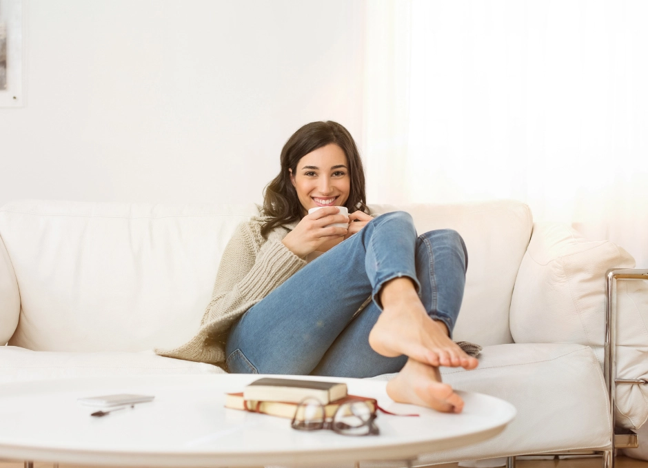 A woman smiling and sitting comfortably on a couch with a book and glasses on the table in front of her, enjoying some stress relief.