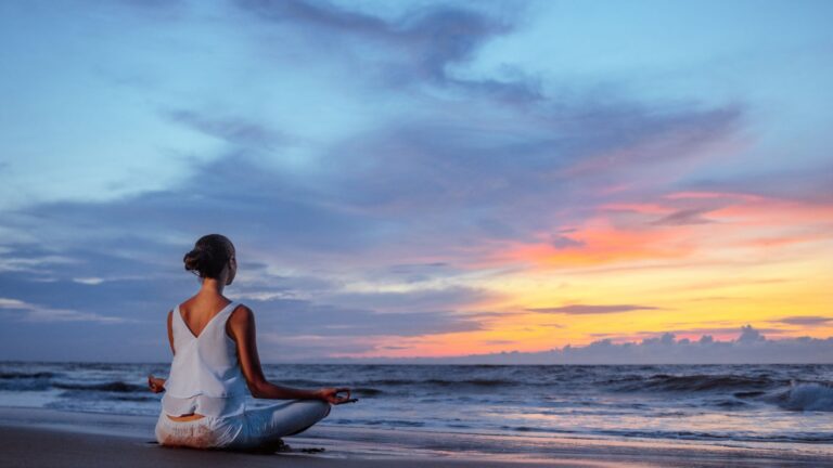 A person sits cross-legged on a beach at sunset, facing the ocean with their hands resting on their knees in a meditative pose.