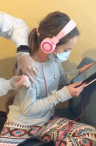 A girl wearing a mask and headphones receives a vaccine injection in her arm while looking at a tablet.