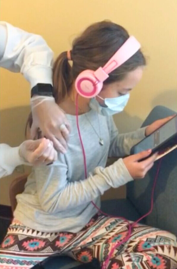 A girl wearing a mask and headphones receives a vaccine injection in her arm while looking at a tablet.