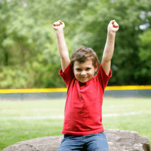 Young child in a red shirt sits on a rock outdoors with both arms raised, smiling—a joyful moment that embodies the calm confidence often encouraged through pediatric hypnosis. Grassy field and trees fill the background.