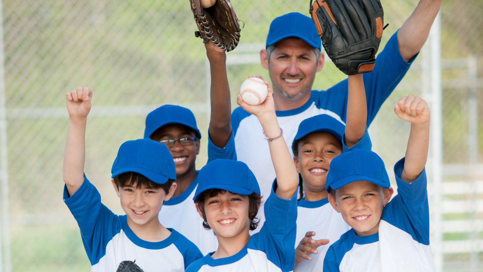 A group of young boys and their coach wearing blue and white baseball uniforms smile and cheer, holding baseball gloves and a ball on a field.